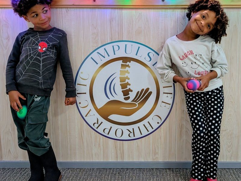 Two children posing next to the Impulse Life Chiropractic logo, decorated with holiday lights